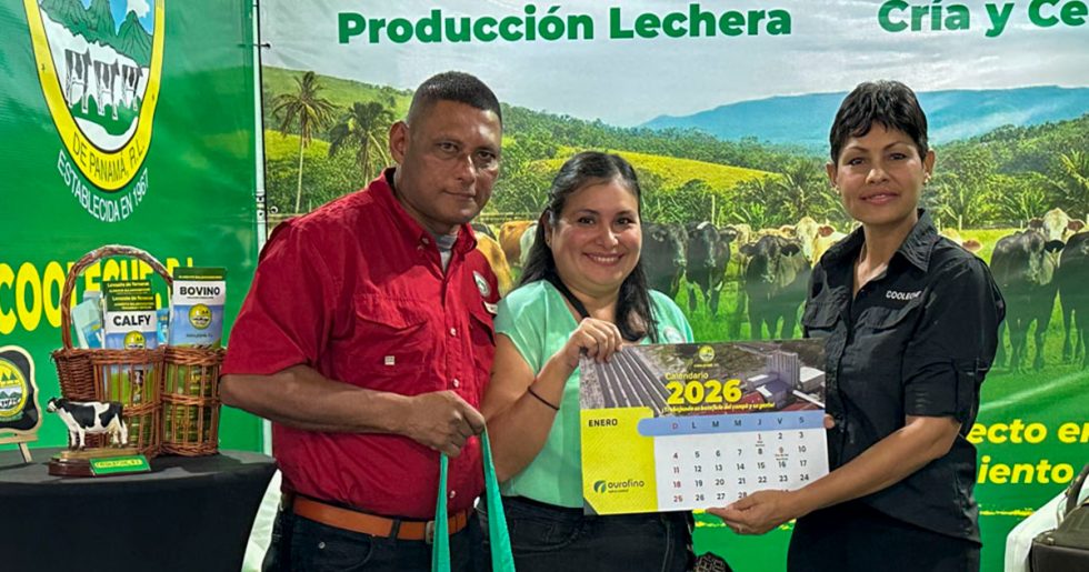 Colaboradora de Cooleche, R.L. entregando un calendario 2026 a dos visitantes en el stand de la cooperativa. De fondo se observa un banner de producción lechera y una exhibición de productos para bovinos.